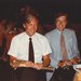 Two men sit at a table in a darkened room and smile at the camera. Other tables and partygoers can be seen sitting at tables in the background.