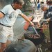 A man in T-shirt and shorts moves meat with a pair of tongs on a smoking charcoal barbecue.