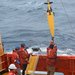 People in bright orange gear stand on a boat in the sea. A yellow oblong shape extends from a rope out of frame.