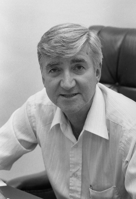 black and white photo of a man with grey hair sitting wearing a white button-up shirt, sitting in a chair