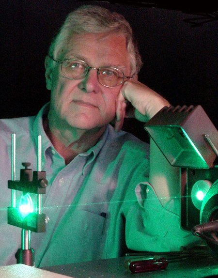 Portrait of an older man with gray hair and glasses posed behind a laser instrument shining green light