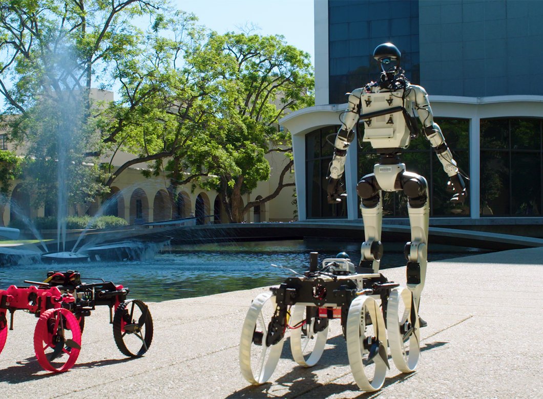 a humanoid robot and two rolling robots in front of Caltech Hall and its fountain at Caltech