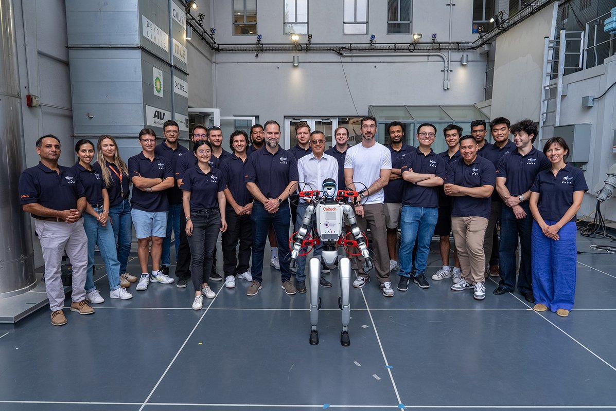 a group of people in CAST wearing CAST/TII collaboration polo shirts, posing with a humanoid robot carrying M4 on its back