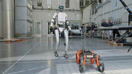 a humanoid robot and a morphing robot in driving mode in CAST with engineers looking on in the background