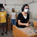 A person in safety glasses and a KN95-style mask holds a sledgehammer poised to smash small rocks on a high, walled table as two other people watch from a few feet back.