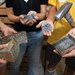 The hands of four people hold rocks in different stages of crushing: whole, sawn into fragments and cleaned and sanded, broken with a sledgehammer (also shown), and crushed in the rock-crusher's tray,