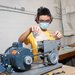 A person in safety glasses and a KN95-style mask feeds rocks from a plastic bag into a crushing machine in a laboratory.