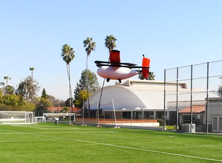 A winged plane with multiple propellers flies above a baseball field in front of Caltech's Brown Gym.