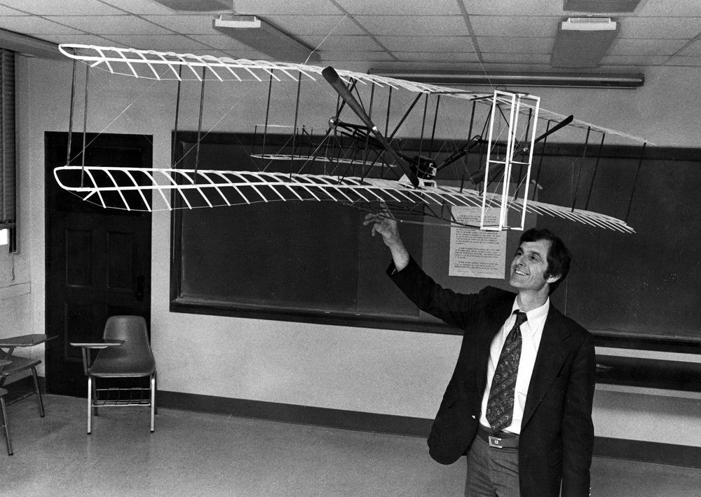 A black and white photo of Fred Culick holding a model of the Wright Flyer over his head in a classroom. A blackboard can be seen behind him.