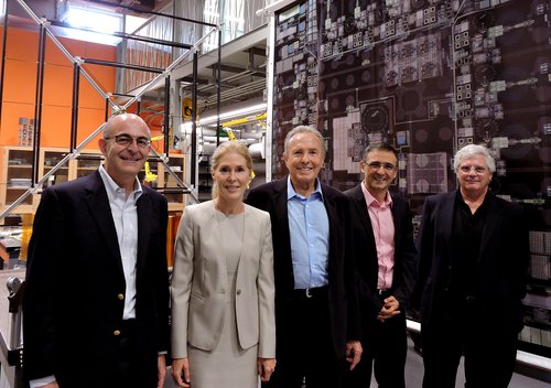 A group photo of five people standing in a high tech laboratory setting. They wear suits and smile at the camera.