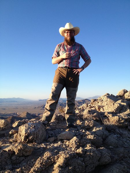 A man wearing a hat and plaid shirt stands among rocks in a dry landscape. Mountains are in the distance