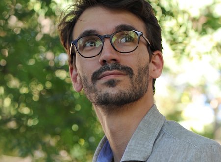 Headshot of a man with dark hair and glasses and a gray collared jacket.