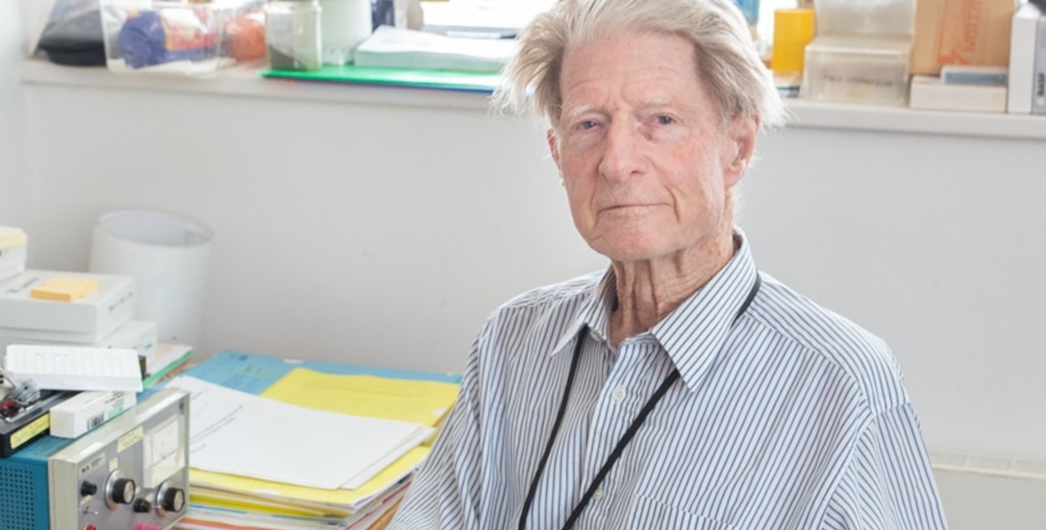 An older man sits at a desk wearing a blue pin-striped shirt.