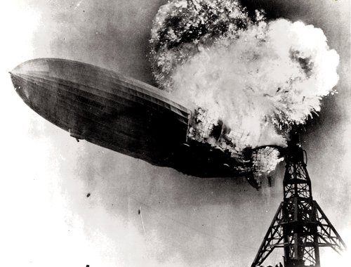 A large zeppelin, partially consumed by flames, begins sinking toward the ground next to its mooring mast. The image is a black and white photograph.