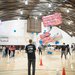 A full scene of the 2024 ME72 competition featuring two teams of students steering sets of white or pink blimps flying above the basketball court in Brown Gym. They are scooping up shiny blue balloons and trying to score them through goals on either end of the court.