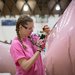 An undergraduate student in a pink shirt uses a screwdriver to make adjustments to the fan on the back of one of her team's blimps.