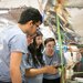 Several students in light blue shirts stare intently at the undercarriage of one of their blimps. One student holds a transmitter that will steer the silver blimp with all of the electrical components shown on the undercarriage.