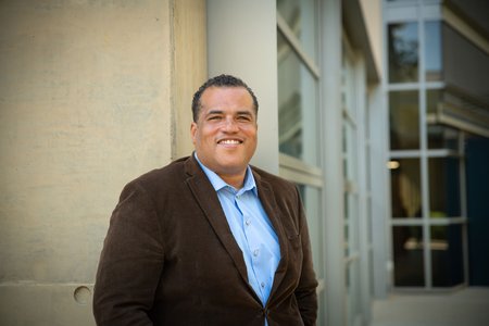 Hosea Nelson poses in front of a campus building, smiling. He wears a button-down shirt and a blazer.