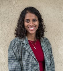 A portrait of Nitika Yadlapalli Yurk. She stands before an outdoor wall, wearing a blazer and smiling.