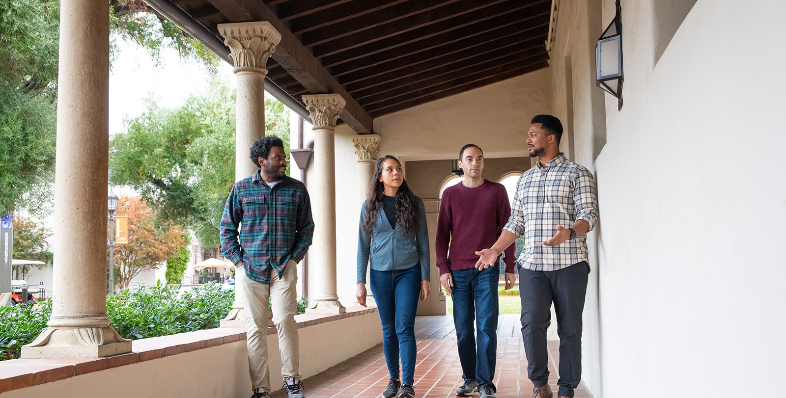 Four scholars talk amongst themselves as they walk alongside a column-lined hallway