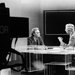 James Quirk sits and converses with an unnamed news anchor at a desk in a television studio. In the foreground, a TV camera labeled "NBC COLOR" can be seen.