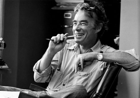 In this black-and-white photo, James Quirk sits casually in a chair, laughing and biting a pencil. He appears to be mid conversation