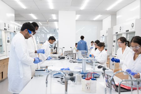 Students in white lab coats along either side of a lab bench in a new lab with fume hoods on either side are engaging in a lab course.