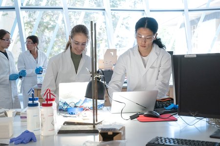 Two students in white lab coats and goggles are working on their laptops at a lab bench with a light on. Two other students are in the background, wearing lap coats and plastic gloves.