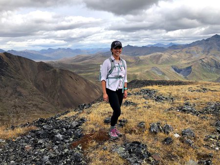 A woman in a hat and hiking gear stands on a hilltop in front of a mountainous landscape.