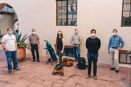 Six people stand outside in masks, posing for a photo next to a large tank of methane and a fan.