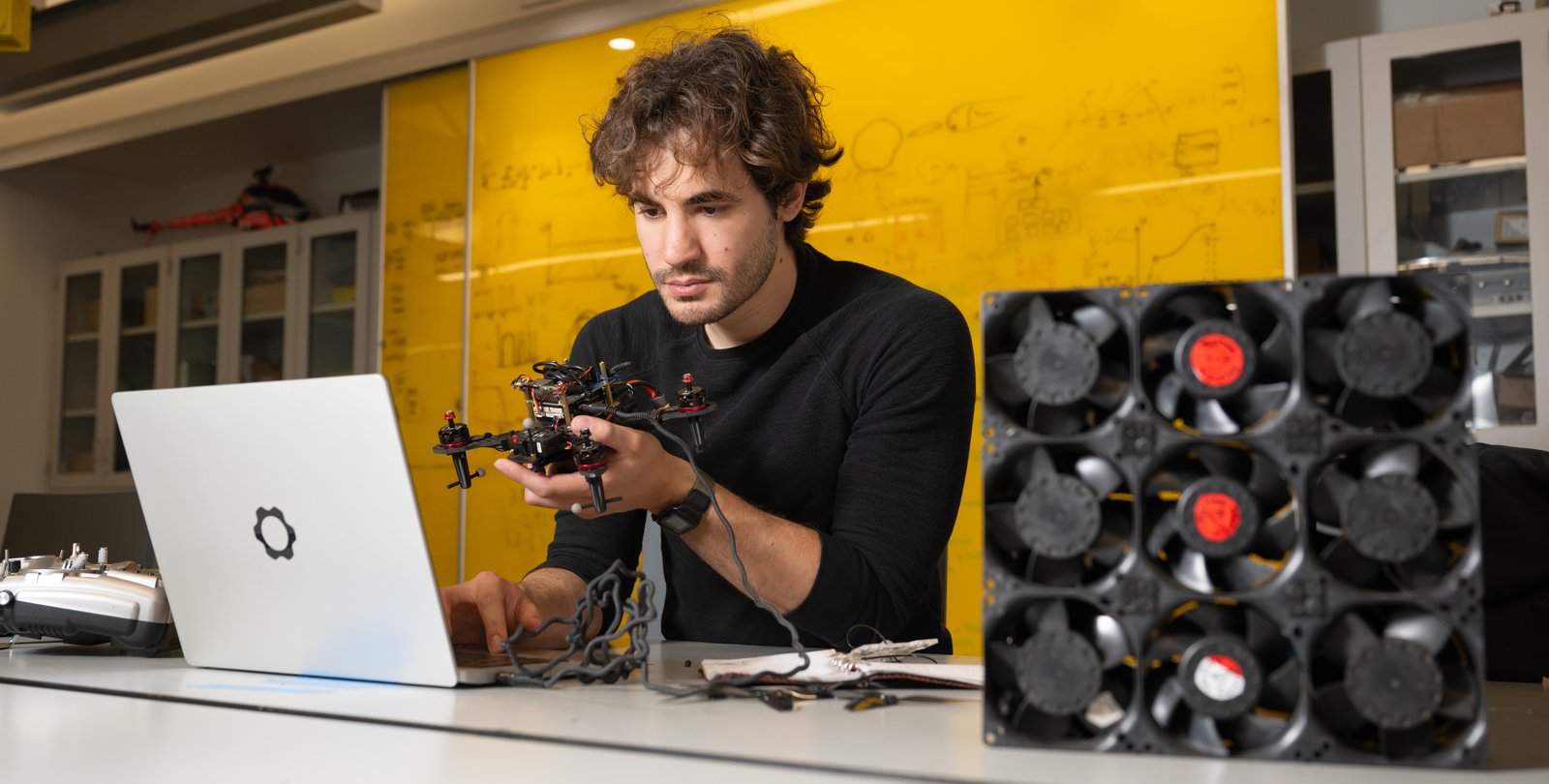 Alejandro Stefan-Zavala sits in front of a laptop with a drone in hand and small fans off to his side.