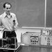 A man standing behind a physics experiment demonstration table in front of a blackboard