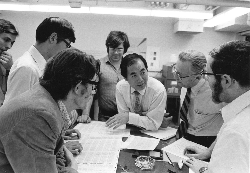 A man wearing a shirt and tie appears at the center of this black and white photo. He leans on a table covered in papers and surrounded by students who are listening to him as he talks.