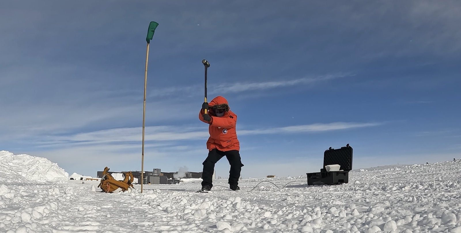 Zhongwen Zhan in a bright red winter coat swinging a sledgehammer at the Antarctic ice
