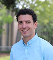 A portrait of Komron Shayegan. He stands outside wearing a button-down shirt and smiling.