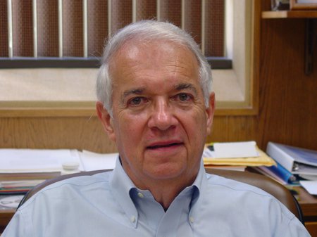 A portrait of Vince McKoy in his office with a window and stacks of paper behind him. He wears a button-down shirt.