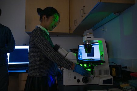 A female graduate student is shown working in a darkened lab space with a fluorescence microscope.